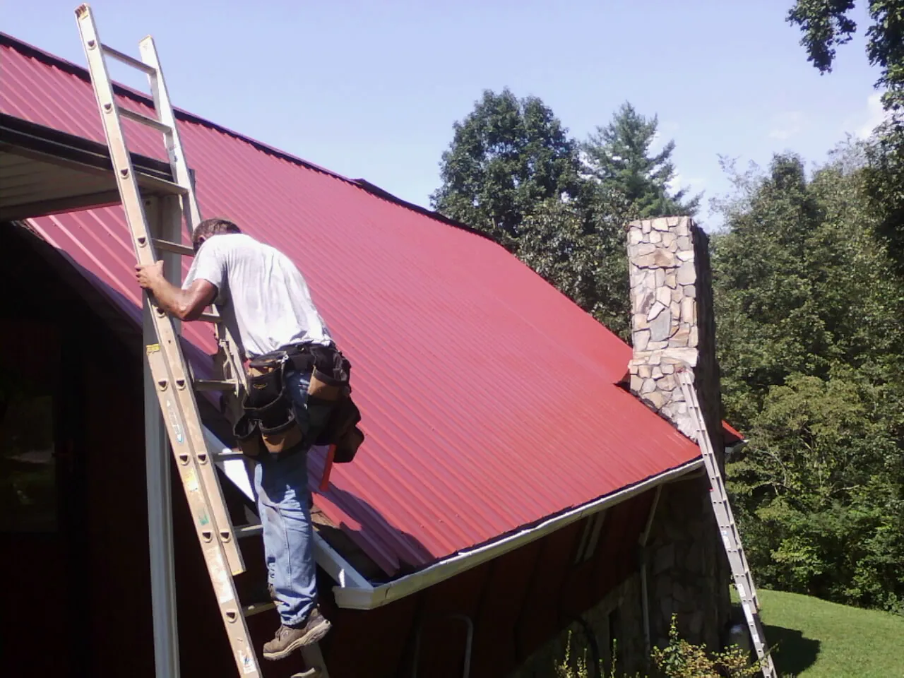 Roofer on a ladder beside a red metal roof and stone chimney — Soffit & Fascia Repair by Roofing Brothers in Woodlawn